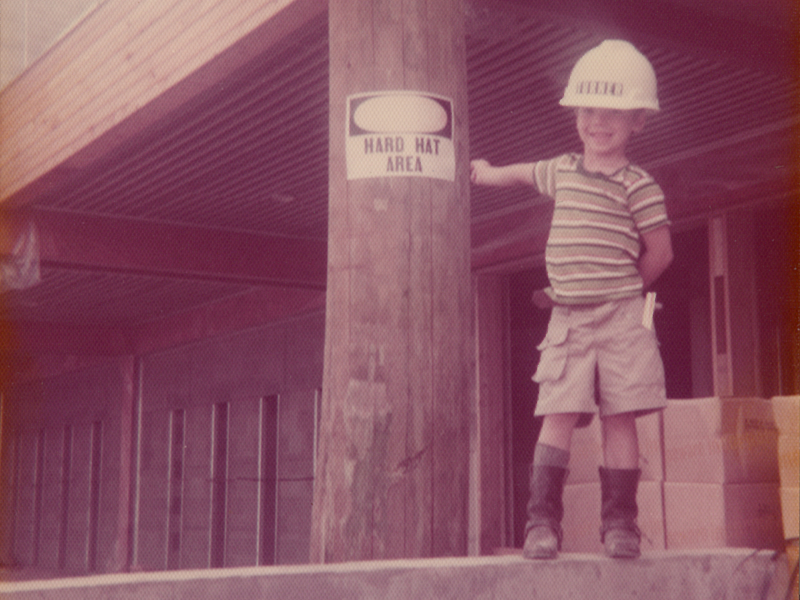A young boy in a striped shirt and hard hat standing on a construction site next to a Hard Hat Area sign, smiling at the camera.